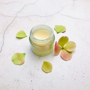 Puede incluir: Un pequeño frasco de vidrio lleno de crema de color amarillo pálido, rodeado de pétalos de flores verdes y rosas sobre un fondo blanco.