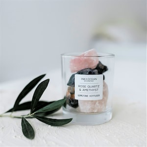 Puede incluir: Un frasco de vidrio lleno de cristales de cuarzo rosa y amatista. El frasco tiene una etiqueta que dice "Ink + Ocean Botanicals Rose Quartz & Amethyst Gemstone Diffuser".