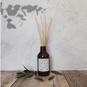Pode incluir: Uma garrafa de vidro marrom com um rótulo preto que diz "INK+OCEAN BOTANICALS ESSENTIAL OIL REED DIFFUSER BASIL TEA TREE GERANIUM NO 3 100ml". A garrafa está cheia de um líquido transparente e tem varetas de madeira que saem do topo. A garrafa está sobre uma superfície de madeira com folhas verdes espalhadas ao redor.