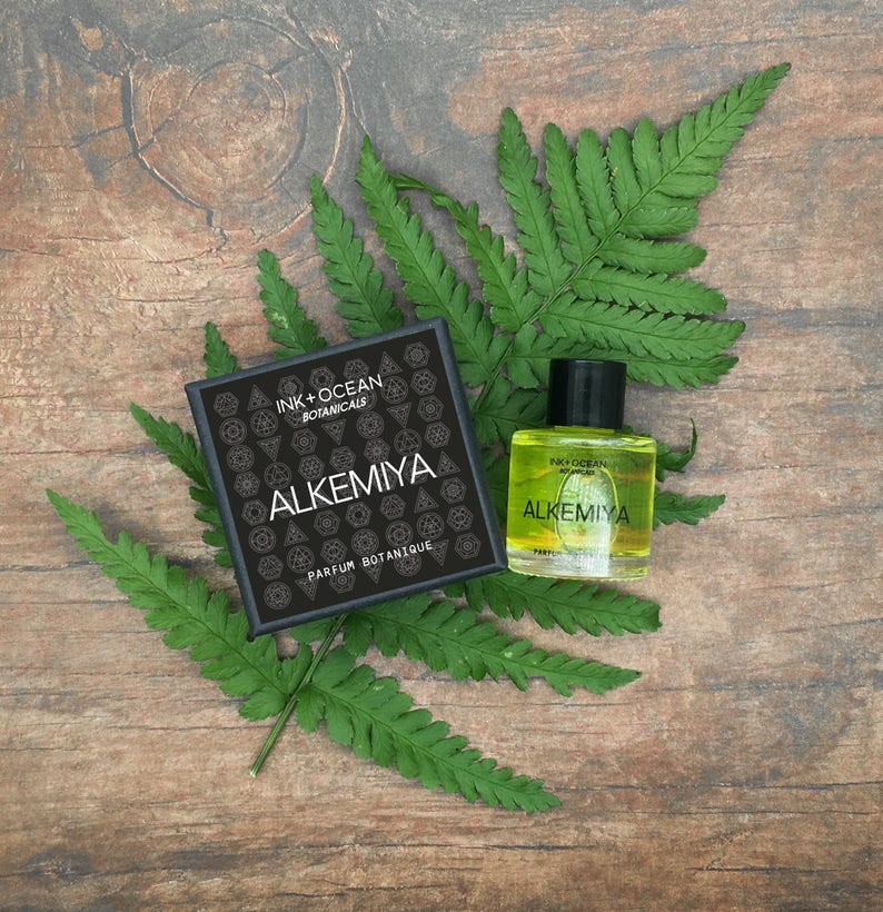 May include: A black box with the text "Ink + Ocean Botanicals Alkemiya Parfum Botanique" and a small bottle of yellow liquid with the same text.