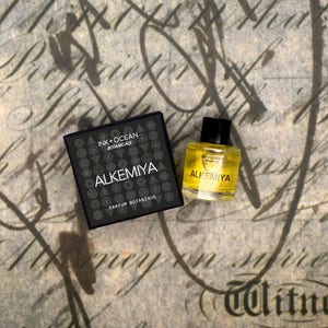 May include: A black box with the text "INK + OCEAN BOTANICALS ALKEMIYA PARFUM BOTANIQUE" and a small bottle of yellow liquid with the text "ALKEMIYA PARFUM BOTANIQUE" on a brown background.