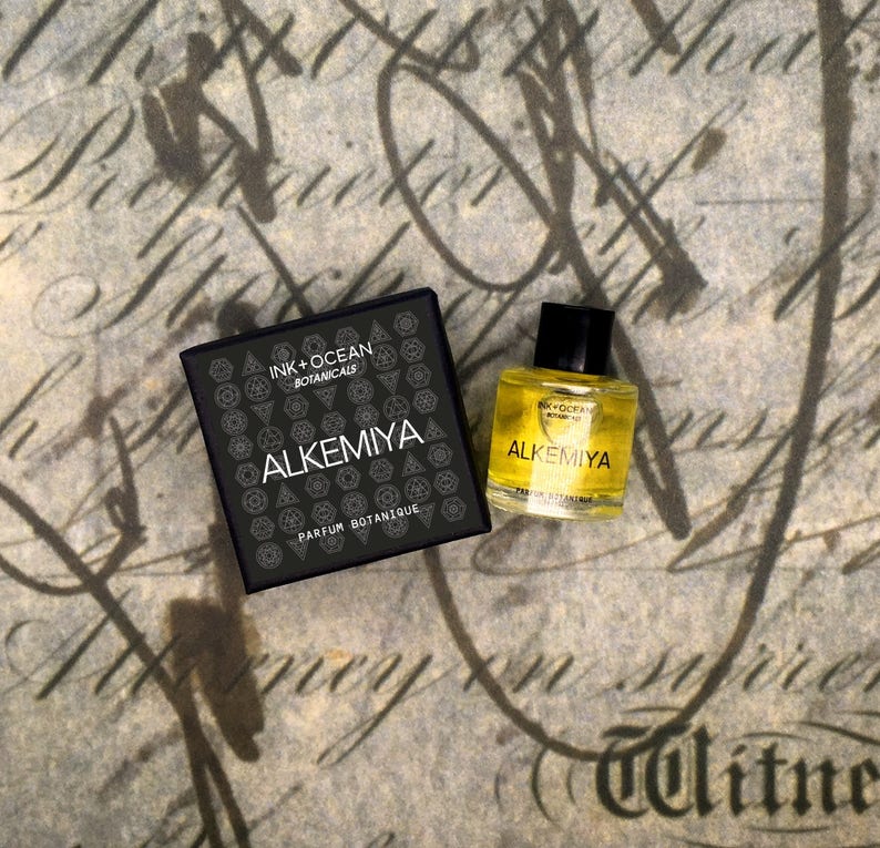 May include: A black box with the text "INK + OCEAN BOTANICALS ALKEMIYA PARFUM BOTANIQUE" and a small bottle of yellow liquid with the text "ALKEMIYA PARFUM BOTANIQUE" on a brown background.