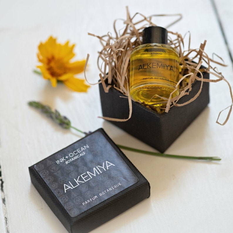 May include: A small black box with a gold label that says "Ink + Ocean Botanicals Alkemiya Parfum Botanique" and a small glass bottle of perfume with a gold label that says "Alkemiya Parfum Botanique".