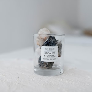 Puede incluir: Un frasco de vidrio lleno de piedras preciosas de sodalita y cuarzo. El frasco tiene una etiqueta que dice "Ink + Ocean Botanicals, Sodalita y Cuarzo, Difusor de piedras preciosas".