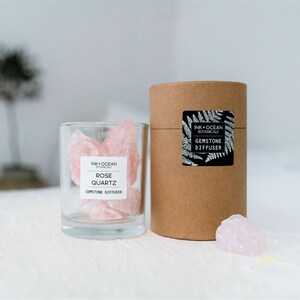 Puede incluir: Un frasco de vidrio lleno de cristales de cuarzo rosa. El frasco tiene una etiqueta que dice "Ink + Ocean Botanicals Rose Quartz Gemstone Diffuser". El frasco está sentado sobre una superficie blanca junto a un tubo de cartón marrón con una etiqueta que dice "Ink + Ocean Botanicals Gemstone Diffuser".