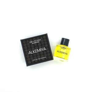 May include: A black box with the text "Ink + Ocean Botanicals" and "Alkemiya Parfum Botanique" on it. A small yellow glass bottle with the same text and a black cap is next to the box.