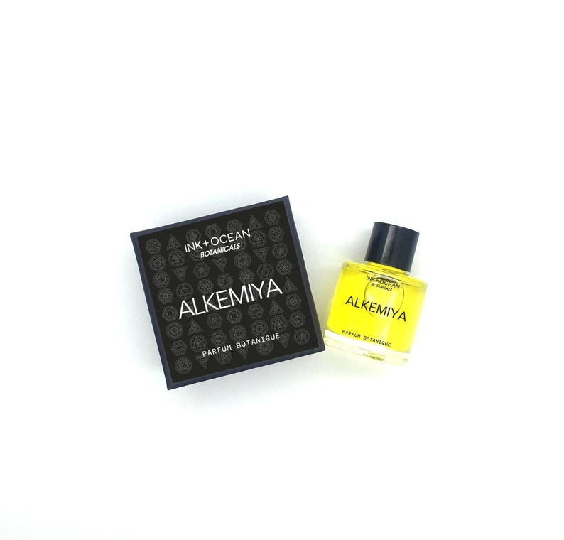 May include: A black box with the text "Ink + Ocean Botanicals" and "Alkemiya Parfum Botanique" on it. A small yellow glass bottle with the same text and a black cap is next to the box.