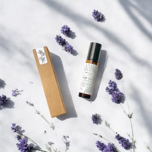 Könnte beinhalten: Eine braune Pappbox und eine Glas-Rollflasche mit schwarzem Verschluss. Die Flasche ist mit "Ink + Ocean" und "And Sleep" sowie einem floralen Design beschriftet. Der Hintergrund ist weiß mit verstreuten Lavendelblüten.