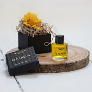 May include: A small bottle of yellow liquid with a black cap and label that reads "Ink + Ocean Botanicals Alkemiya Parfum Botanique". The bottle is sitting on a wooden round slice. A small black box with a yellow flower on top is in the background.