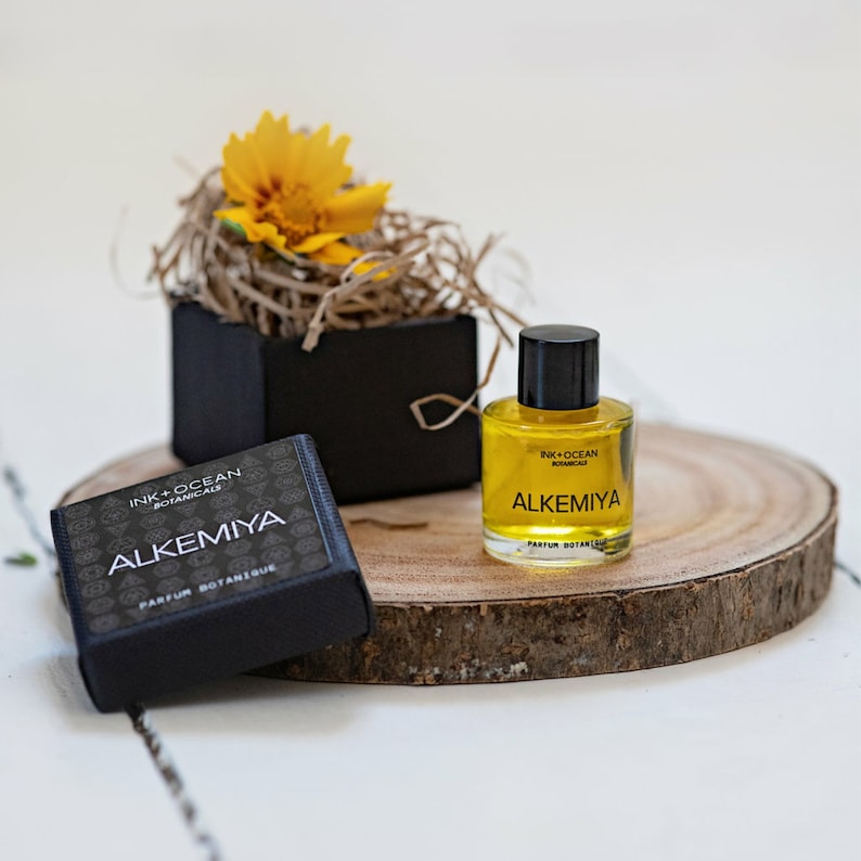 May include: A small bottle of yellow liquid with a black cap and label that reads "Ink + Ocean Botanicals Alkemiya Parfum Botanique". The bottle is sitting on a wooden round slice. A small black box with a yellow flower on top is in the background.