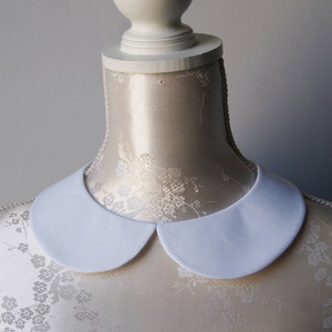 May include: A white, rounded Peter Pan collar displayed on a mannequin. The collar is made of a smooth, opaque fabric and is a classic style. The mannequin is a light beige color.