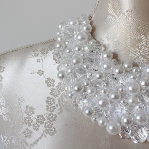 May include: A white beaded necklace with faux pearls and clear crystal accents. The necklace is displayed on a mannequin with a floral patterned fabric. The pearls vary in size, creating a textured, layered effect. This statement necklace is perfect for special occasions.