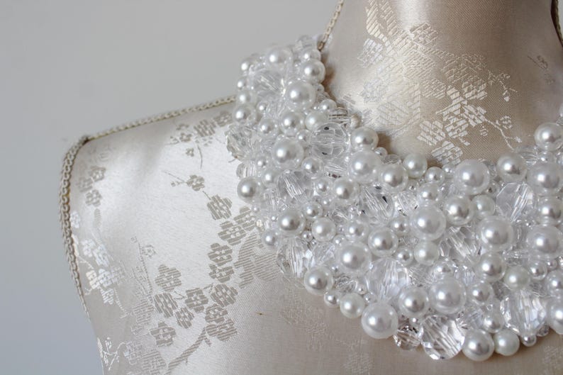 May include: A white beaded necklace with faux pearls and clear crystal accents. The necklace is displayed on a mannequin with a floral patterned fabric. The pearls vary in size, creating a textured, layered effect. This statement necklace is perfect for special occasions.