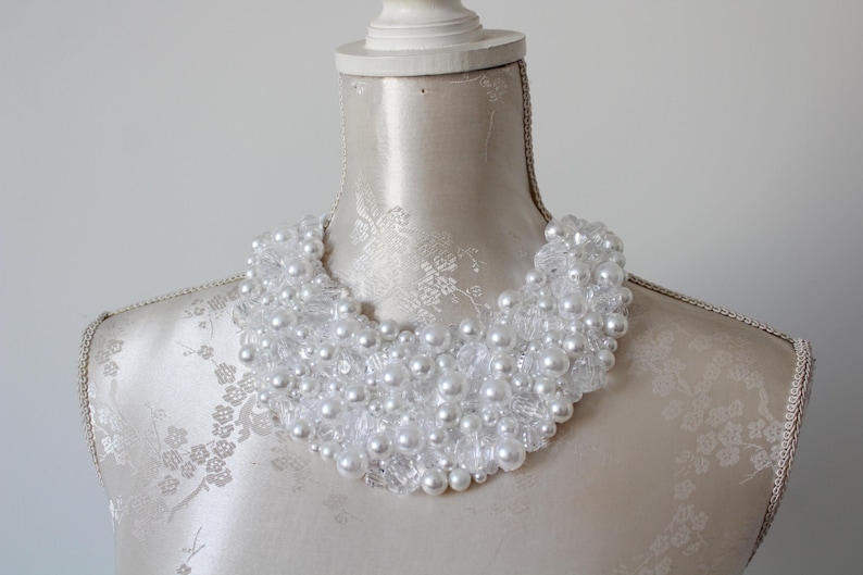 May include: A white beaded collar necklace on a mannequin. The necklace features a mix of clear, faceted beads and larger, round, white pearls. The beads are densely arranged, creating a statement piece. The mannequin is a neutral color.