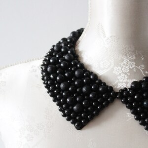 May include: A black beaded collar necklace displayed on a white mannequin. The collar is made of various sized black beads, creating a textured, layered effect. The collar is a statement piece, perfect for adding a touch of elegance.