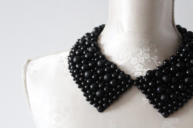 May include: A black beaded collar necklace displayed on a white mannequin. The collar is made of various sized black beads, creating a textured, layered effect. The collar is a statement piece, perfect for adding a touch of elegance.