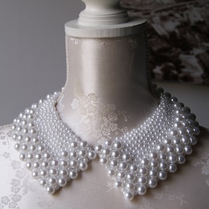 Può includere: Una collana di perle bianche a forma di colletto. La collana è composta da molte piccole perle ed è indossata su un manichino bianco.