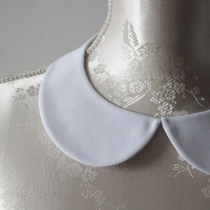 May include: A white, rounded Peter Pan collar, likely for a blouse or dress, is displayed on a mannequin. The collar has a smooth, matte finish and a classic, timeless design. The background is a soft, neutral color, highlighting the collar's clean lines.