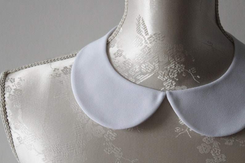 May include: A white, rounded Peter Pan collar, likely for a blouse or dress, is displayed on a mannequin. The collar has a smooth, matte finish and a classic, timeless design. The background is a soft, neutral color, highlighting the collar's clean lines.