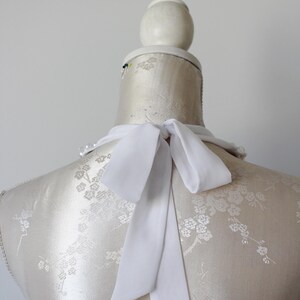 May include: A white silk necktie is tied in a bow on a mannequin. The mannequin is covered in a cream-colored fabric with a floral pattern. The background is a plain, light gray.