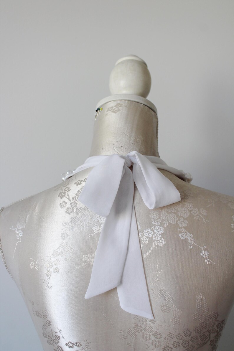 May include: A white silk necktie is tied in a bow on a mannequin. The mannequin is covered in a cream-colored fabric with a floral pattern. The background is a plain, light gray.