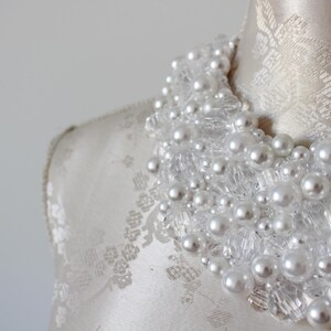 May include: A close-up of a white beaded necklace with faux pearls and clear crystal accents. The necklace is displayed on a mannequin with a cream-colored fabric. The beads are clustered together.