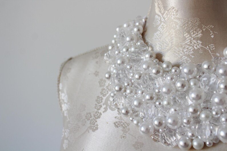 May include: A close-up of a white beaded necklace with faux pearls and clear crystal accents. The necklace is displayed on a mannequin with a cream-colored fabric. The beads are clustered together.