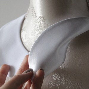 May include: A white, satin-like collar, likely for a shirt or blouse, is displayed on a mannequin. The collar has a rounded shape and is being held by a hand. The background is a neutral color.