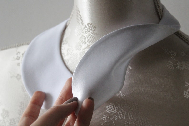 May include: A white, satin-like collar, likely for a shirt or blouse, is displayed on a mannequin. The collar has a rounded shape and is being held by a hand. The background is a neutral color.