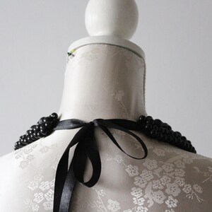 May include: A black beaded necklace with a satin ribbon tie, displayed on a white mannequin. The necklace features multiple strands of small, round beads. The ribbon is tied in a bow at the back.