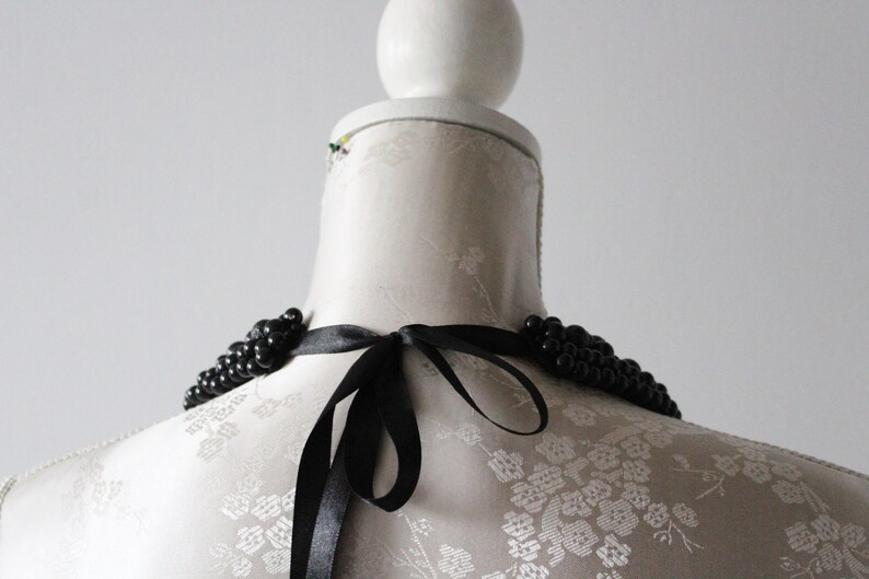 May include: A black beaded necklace with a satin ribbon tie, displayed on a white mannequin. The necklace features multiple strands of small, round beads. The ribbon is tied in a bow at the back.