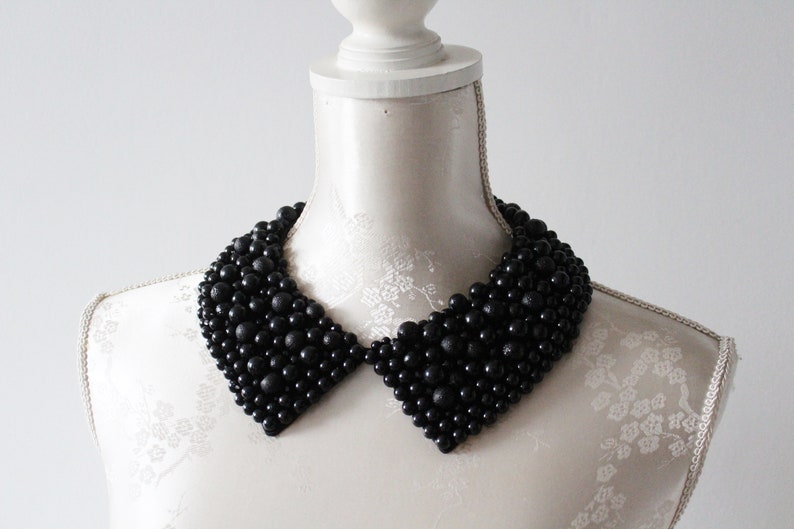 May include: A black beaded collar necklace displayed on a white mannequin. The collar is made of numerous small, round, black beads, creating a textured, layered effect. The collar has a classic, pointed shape, and is a statement piece.