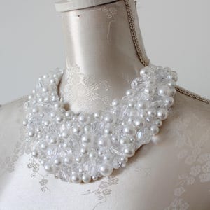 May include: A white beaded necklace with large and small faux pearls and clear crystals. The necklace is displayed on a mannequin with a cream-colored fabric. The necklace is a statement piece.