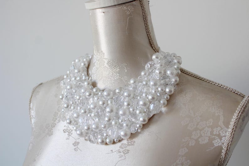 May include: A white beaded necklace with large and small faux pearls and clear crystals. The necklace is displayed on a mannequin with a cream-colored fabric. The necklace is a statement piece.