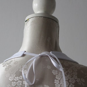 May include: A white detachable collar with a ribbon tie, displayed on a mannequin. The collar has a classic, pointed shape and is made of a smooth, opaque fabric. The mannequin has a floral pattern.