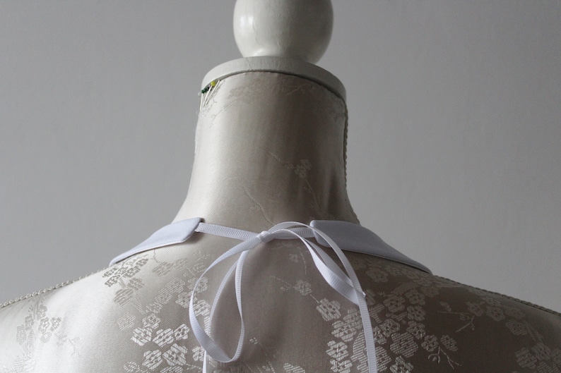 May include: A white detachable collar with a ribbon tie, displayed on a mannequin. The collar has a classic, pointed shape and is made of a smooth, opaque fabric. The mannequin has a floral pattern.