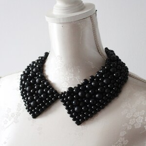 May include: A black beaded collar necklace displayed on a white mannequin. The collar is made of numerous small, round, black beads, creating a textured surface. The collar has a classic, pointed shape, and is a statement piece of jewelry.