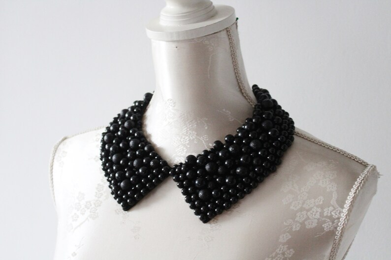 May include: A black beaded collar necklace displayed on a white mannequin. The collar is made of numerous small, round, black beads, creating a textured surface. The collar has a classic, pointed shape, and is a statement piece of jewelry.