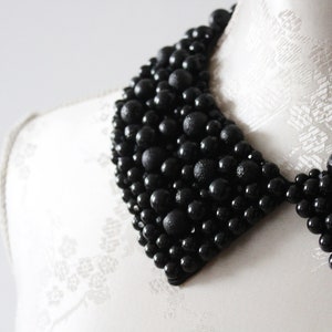 May include: A black beaded collar necklace. The collar is made of various sizes of black beads, creating a textured and dimensional effect. The necklace is displayed on a white mannequin.
