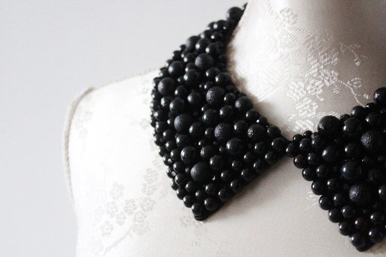 May include: A black beaded collar necklace. The collar is made of various sizes of black beads, creating a textured and dimensional effect. The necklace is displayed on a white mannequin.