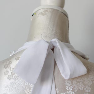 May include: A white silk scarf tied in a bow around the neck of a mannequin. The mannequin is covered in a floral pattern. The scarf is made of a lightweight, flowing fabric.