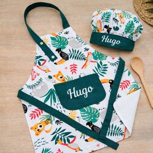 Puede incluir: Un delantal blanco y un gorro de chef a juego con un colorido estampado tropical de animales y hojas. El delantal tiene un bolsillo y una cinta para el cuello de color verde oscuro, ambos bordados con el nombre Hugo en blanco. El gorro también lleva el nombre Hugo.