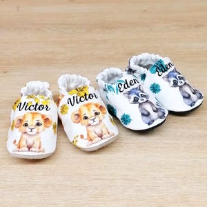 Peut inclure: Quatre paires de chaussures pour bébés avec des motifs d'animaux. Deux paires présentent un lion avec le nom "Victor", et les deux autres paires ont un raton laveur avec le nom "Eden". Les chaussures sont blanches avec des accents colorés et une semelle souple.