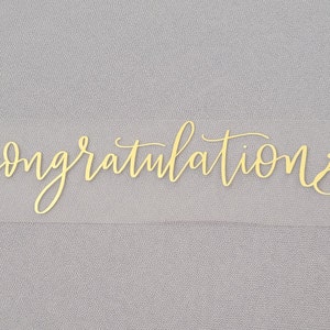 May include: A clear sticker with the word "congratulations" written in gold cursive lettering.