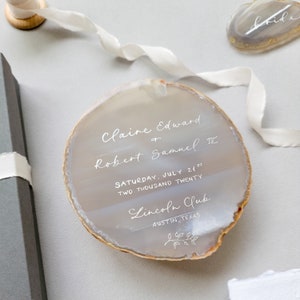 Silver calligraphy agate slice wedding invitation, save the date, on a light gray/ silver and tan natural stone,  spill off silk ribbon, box, and agate spice place cards in the background