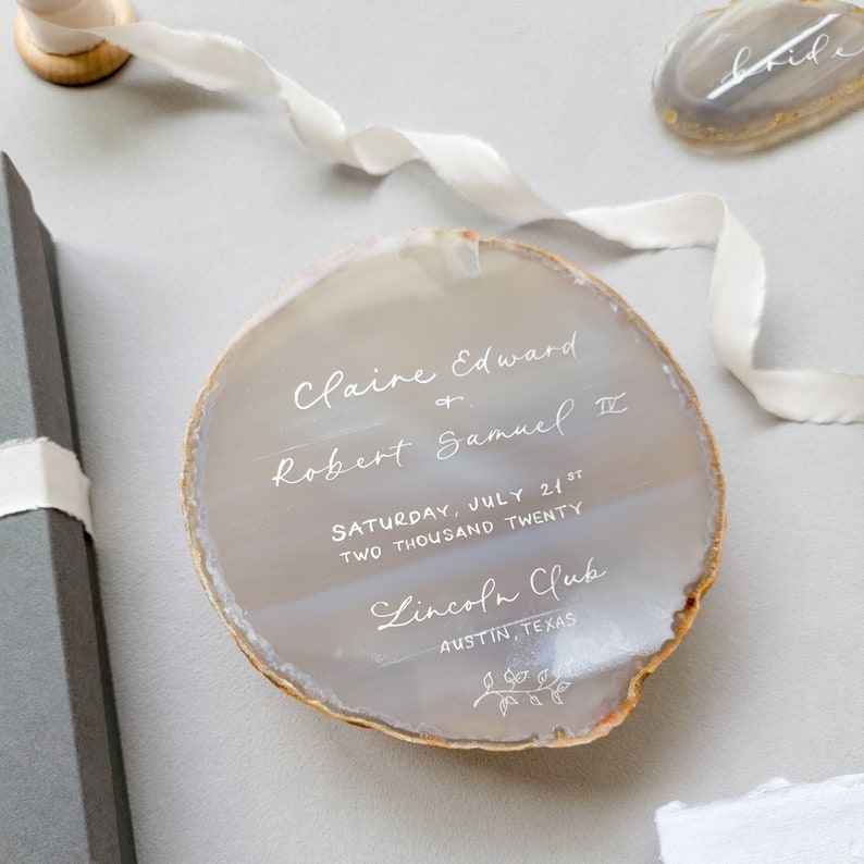 Silver calligraphy agate slice wedding invitation, save the date, on a light gray/ silver and tan natural stone,  spill off silk ribbon, box, and agate spice place cards in the background