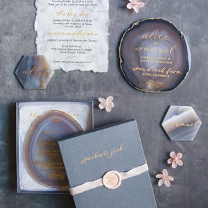 Gold hand calligraphy on a neutral gray blue agate stone (charcoal gray boxed wedding invitation) on a charcoal background and decorated with cherry blossom flowers and two hexagon agates place cards with gold calligraphy