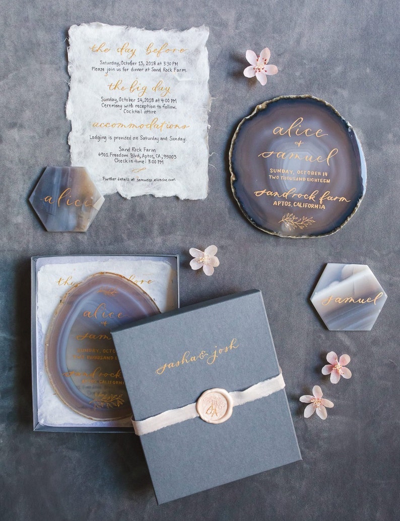 Gold hand calligraphy on a neutral gray blue agate stone (charcoal gray boxed wedding invitation) on a charcoal background and decorated with cherry blossom flowers and two hexagon agates place cards with gold calligraphy
