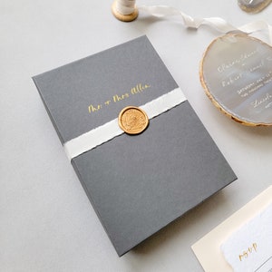 Personalized charcoal gray boxed wedding invitation, gold calligraphy by Jen, wrapped with a gold wax seal and cream hand-dyed silk ribbon, agate invitation and rsvp with envelope on a light gray background