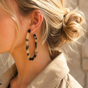 May include: A large oval hoop earring featuring a smooth, polished finish with a distinctive tortoiseshell pattern in shades of cream, tan, and dark brown. The earring has a slender, rounded profile and is designed to hang vertically from the earlobe.